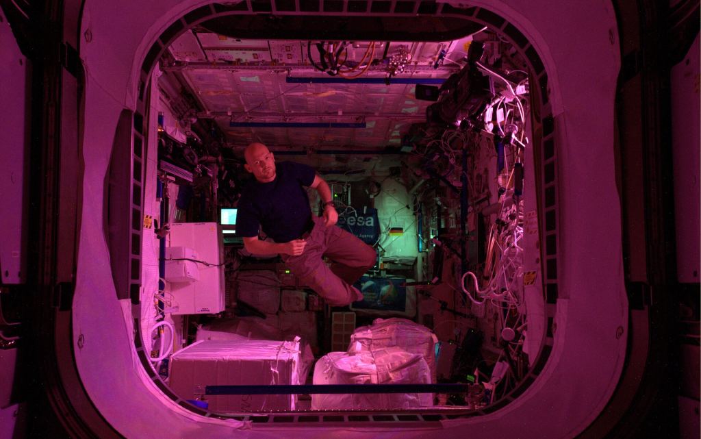 Astronauta che galleggia nel modulo Columbus immerso in una morbida luce rosa.
